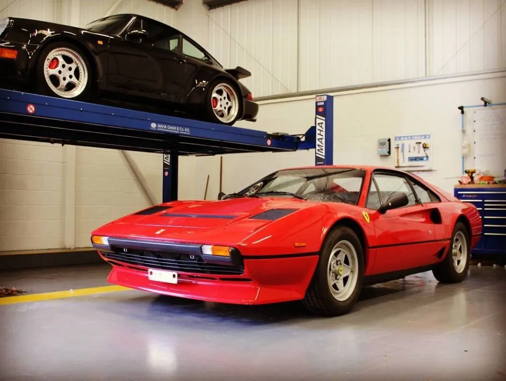 A classic red Ferrari 308 in a workshop, with a Porsche 911 on the lift above